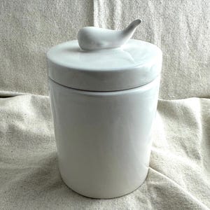 May include: A white ceramic jar with a lid and a decorative handle. The jar is cylindrical and sits on a neutral-colored fabric. The lid has a curved handle. The overall design is minimalist and modern.