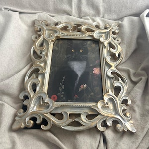 May include: An ornate silver picture frame featuring a portrait of a black cat with yellow eyes and a white chest. The cat is set against a floral backdrop with red and pink flowers. The frame has a detailed, scrollwork design.