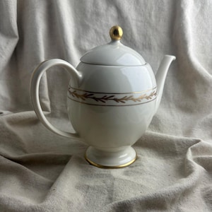 May include: A white porcelain teapot featuring a rounded body, a curved handle, and a spout. The lid is topped with a gold-colored knob. A horizontal band with a gold leaf-patterned design encircles the center, and the base has a gold rim.
