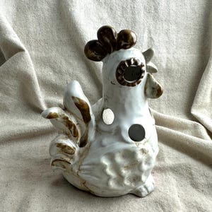 May include: A white ceramic rooster figurine featuring brown painted accents on the comb, eye area, beak, and tail feathers. The body of the rooster has two circular cutouts on the side, and the surface has a textured, glossy finish.