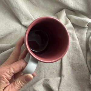 May include: A ceramic mug with a white handle and a dark pink interior. The exterior of the mug is a lighter pink. The mug is empty and appears to be clean.