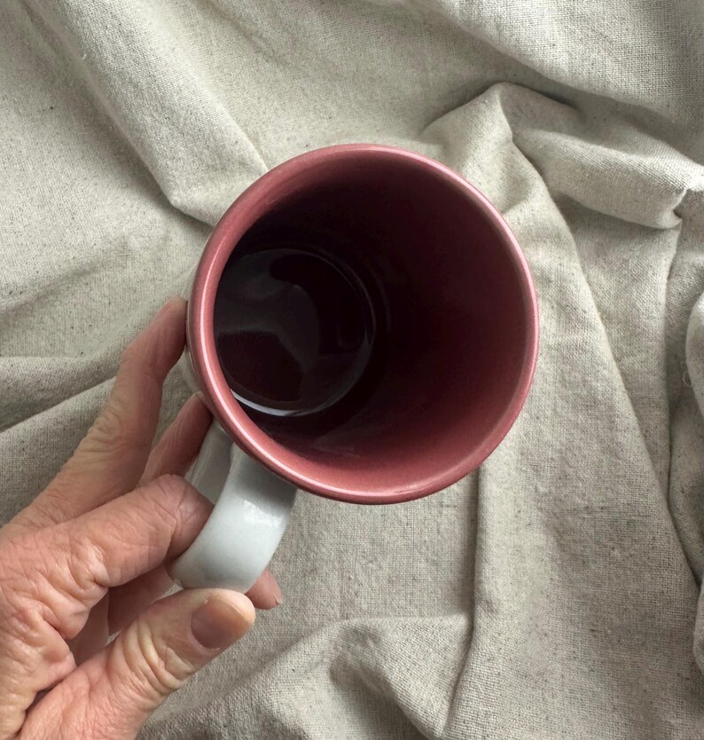 May include: A ceramic mug with a white handle and a dark pink interior. The exterior of the mug is a lighter pink. The mug is empty and appears to be clean.