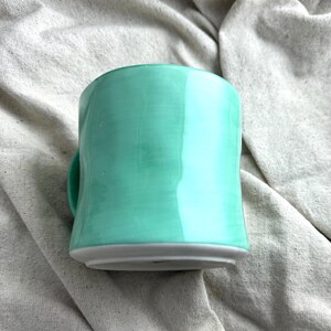May include: A handmade ceramic mug in a vibrant turquoise glaze. The mug has a rounded shape with a small handle and a gray base. The mug is sitting on a neutral-colored fabric.