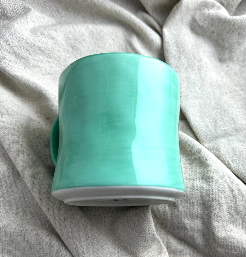 May include: A handmade ceramic mug in a vibrant turquoise glaze. The mug has a rounded shape with a small handle and a gray base. The mug is sitting on a neutral-colored fabric.