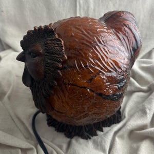 May include: A detailed, brown rooster figurine with a glossy finish. The sculpture features intricate detailing of feathers, a comb, and a wattle. A black electrical cord extends from the base. The rooster is a decorative item.