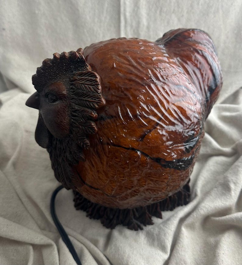 May include: A detailed, brown rooster figurine with a glossy finish. The sculpture features intricate detailing of feathers, a comb, and a wattle. A black electrical cord extends from the base. The rooster is a decorative item.