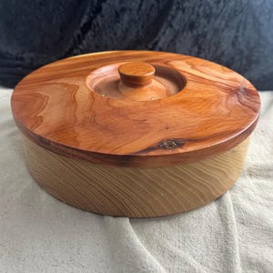 May include: A round wooden box with a lid. The box is made of light wood with a darker wood lid. The lid has a small knob on top.