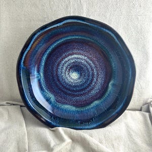 May include: A handmade ceramic plate with a unique, organic shape. The plate features a glossy, deep blue glaze with swirling patterns of lighter blues, purples, and hints of brown, creating a visually appealing, artistic design. The plate's rim has an uneven edge.
