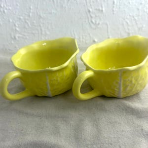 May include: Two bright yellow ceramic cups with handles. The cups have a textured, cabbage-like design on the exterior and a smooth interior. The cups are sitting on a light-colored surface.