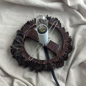 May include: A vintage-style lamp with a dark brown, ornate, circular frame. The frame surrounds a white light socket holding a small light bulb. The lamp has a black power cord and is set against a neutral fabric background.