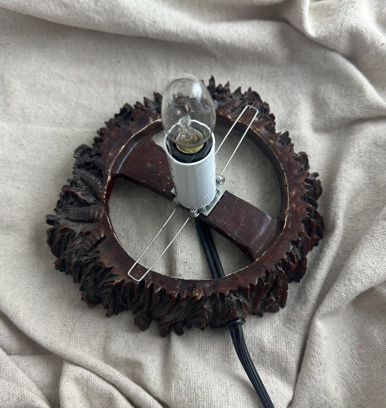 May include: A vintage-style lamp with a dark brown, ornate, circular frame. The frame surrounds a white light socket holding a small light bulb. The lamp has a black power cord and is set against a neutral fabric background.