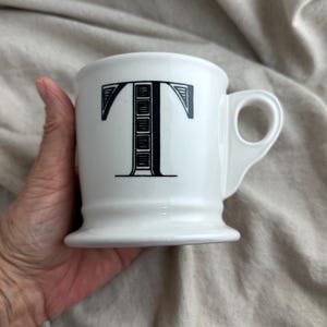 May include: White ceramic mug with a black letter "T" design. The mug has a rounded base and a handle. The mug is held in a hand.