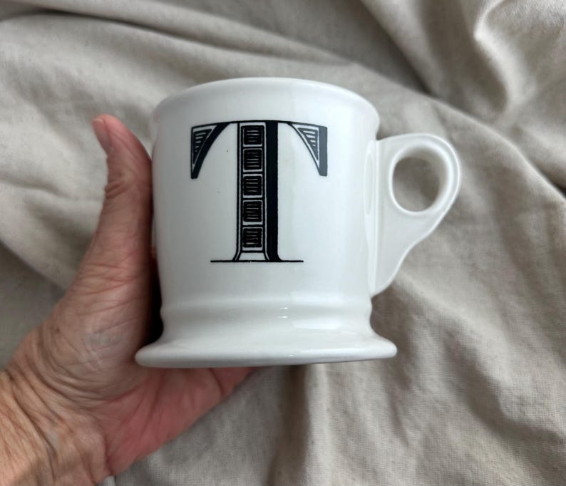 May include: White ceramic mug with a black letter "T" design. The mug has a rounded base and a handle. The mug is held in a hand.