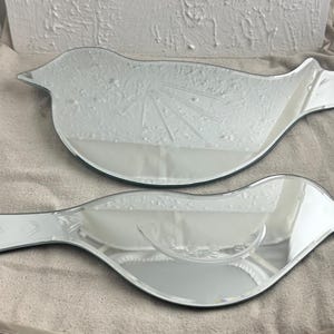 May include: Two silver bird-shaped mirrors with a decorative etched design. The mirrors are on a white fabric surface.