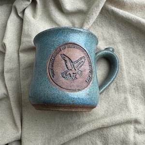 May include: A blue-green ceramic mug with a speckled finish and a brown base. A circular medallion on the front features an embossed eagle graphic and the text "THEY SHALL MOUNT UP WITH WINGS AS EAGLES IS 40:31" encircling the bird in raised lettering.