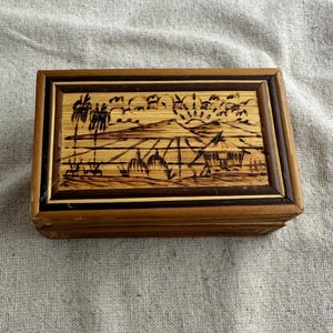 May include: A rectangular wooden box with a detailed landscape scene on the lid. The scene depicts palm trees, huts, and a sunrise, all in a dark brown color. The box has a dark brown border and a light brown frame.