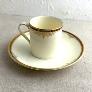 May include: A white porcelain teacup and saucer with a gold and burgundy trim. The cup and saucer have a delicate floral design in gold.