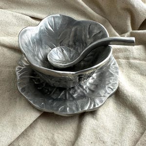 May include: A decorative silver-colored serving set, including a bowl, a small spoon, and a saucer, all with a leaf-like design. The bowl and saucer are approximately 6 inches in diameter.