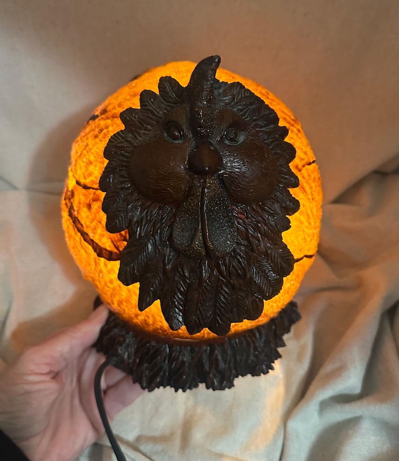 May include: A decorative lamp shaped like a rooster's head. The lamp has a round, orange-colored body with a textured surface. The rooster's head is dark brown with detailed features. The lamp is lit from within, creating a warm glow.