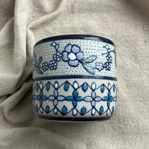 May include: A cylindrical ceramic mug featuring a cream-colored base with two distinct horizontal bands. The upper band displays a delicate floral pattern in shades of blue and green on a dotted background, while the lower band features a geometric lattice design in blue and white.