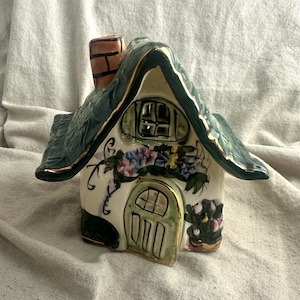 May include: A ceramic house-shaped figurine featuring a dark green textured roof with gold trim, a light-colored facade, a green arched door, a chimney, and painted floral details including blue and pink flowers and green foliage accents.