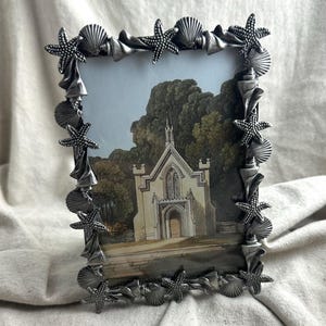 May include: A rectangular picture frame featuring a metallic silver finish with a decorative border composed of sculpted starfish and various seashells. The frame holds an illustration of a stone building with arched windows and doors.