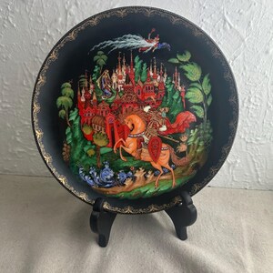 May include: A black and gold decorative plate with a painted scene of a knight riding a horse through a forest. The scene includes a castle in the background and a flying figure in the sky.