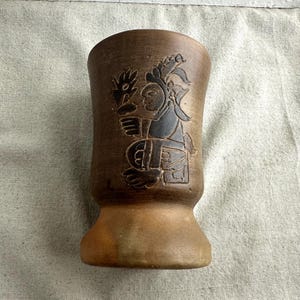 May include: A brown ceramic vase with a flared base and a cylindrical body. The vase features a carved design of a figure holding a flower. The figure is depicted in a traditional style, with intricate details. The vase has a rustic, handcrafted appearance.