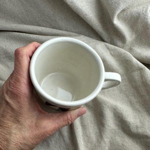 May include: A white ceramic mug with a handle, held in a hand. The mug has a simple design and a smooth, glossy finish. The interior of the mug is white, and the handle is curved. The mug is held against a neutral-colored fabric background.