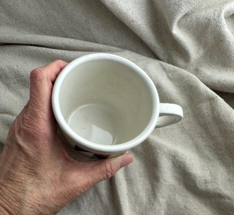 May include: A white ceramic mug with a handle, held in a hand. The mug has a simple design and a smooth, glossy finish. The interior of the mug is white, and the handle is curved. The mug is held against a neutral-colored fabric background.