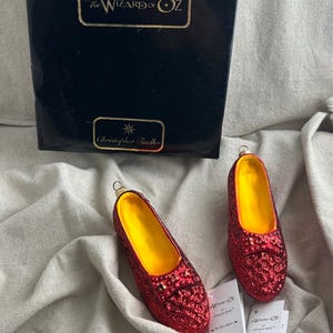 May include: Two red glittery shoe ornaments with yellow interiors, inspired by The Wizard of Oz, sit next to a black box with gold lettering. The box reads "The Wizard of Oz" and "Christopher Radko".