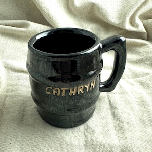 May include: A black ceramic mug with a barrel-like design and a handle. The mug has the name "CATHRYN" in gold lettering. The mug is sitting on a beige fabric surface.