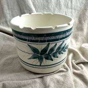 May include: A cream-coloured ceramic bowl with a wide, notched rim. The exterior features a horizontal band of dark green and light purple stripes, with a hand-painted green leaf motif and a thin, cursive script encircling the upper section.