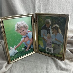 May include: A gold-colored, hinged picture frame with two panels. Each panel holds a photograph. One photo shows a child with a rabbit, and the other shows a family. The frame is labeled "5x7".
