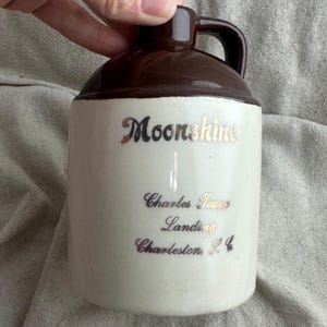 May include: A vintage ceramic jug with a brown top and handle, and a cream-colored body. The jug has the word "Moonshine" in silver script, along with "Charles Towne Landing, Charleston, SC."