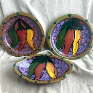 May include: Two round ceramic plates and one oval ceramic bowl featuring a hand-painted design of three chili peppers in green, red, and yellow. The background of each piece is a light purple speckled pattern with a tan border decorated with green dots.