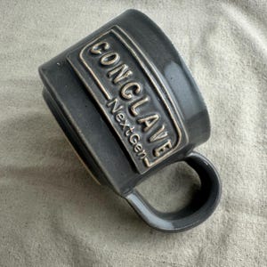 May include: A dark gray ceramic mug with a rounded handle. The mug features the word "CONCLAVE" in raised lettering within a rectangular frame, with "NextGen" below. The mug is angled, showcasing its design.