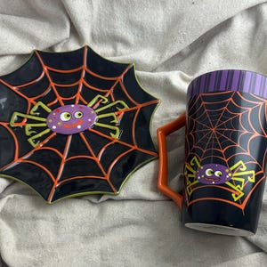 May include: A Halloween-themed ceramic mug and plate set. The black mug and plate feature orange spiderwebs and cartoon spiders. The mug has a purple striped rim and an orange handle. The plate is shaped like a spiderweb.