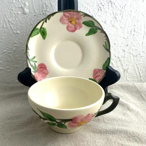 May include: Cream-colored teacup and saucer set with pink floral and green leaf design. The teacup has a dark handle, and the saucer has a scalloped edge. The set is displayed on a neutral-colored surface.