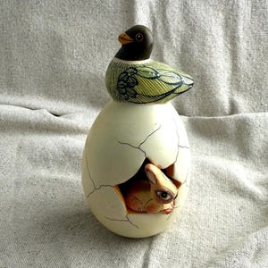May include: A whimsical ceramic figurine featuring a duck perched atop a cracked egg, with a rabbit peeking out. The duck has a black head and green and yellow wings. The egg is off-white with brown cracks, and the rabbit is tan.