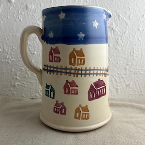 May include: A white ceramic pitcher with a blue and white star pattern on the top. The pitcher has a handle and is decorated with a colorful pattern of houses.