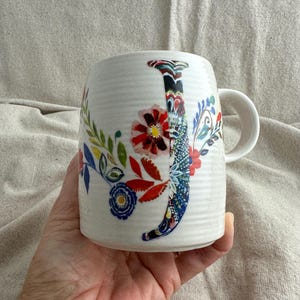 Op de afbeelding: Witte keramische mok met een bloemenontwerp en de letter "J". De mok heeft een gebogen handvat en is versierd met kleurrijke bloemen, bladeren en een patroonletter. De mok wordt in een hand gehouden.