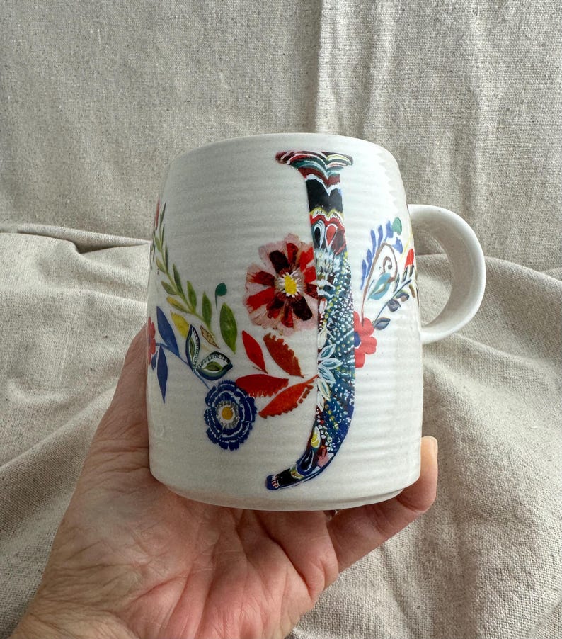 Op de afbeelding: Witte keramische mok met een bloemenontwerp en de letter "J". De mok heeft een gebogen handvat en is versierd met kleurrijke bloemen, bladeren en een patroonletter. De mok wordt in een hand gehouden.