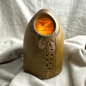 May include: A brown ceramic candle holder shaped like a hooded figure with a rounded top. The front features a circular opening revealing a warm orange glow, carved hands held together, a vertical row of small circular cutouts, and a cross symbol.