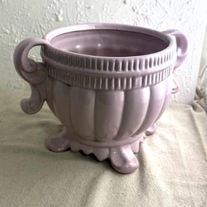 May include: A light purple ceramic planter with two curved handles and a scalloped base. The planter has a ribbed design on the body and a decorative band near the rim. The planter is a decorative item.