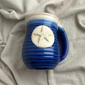 May include: A ceramic mug featuring a deep blue glazed finish with horizontal ribbed textures. The mug has a prominent circular cream-coloured medallion on the front displaying a raised, textured starfish relief and a smooth cream-coloured rim.