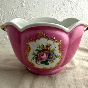 May include: A pink ceramic planter with a scalloped rim and floral design. The planter features a white central panel with a bouquet of colorful flowers, accented by gold trim. The exterior is a glossy pink.