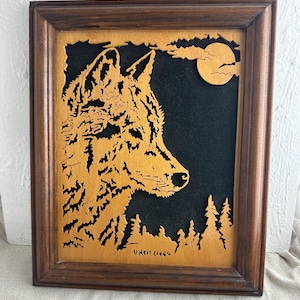 May include: Wooden framed art piece featuring a wolf profile, moon, and trees. The wolf is cut from light-colored wood, set against a black background. The frame is a dark brown color. The artwork is signed "UNO! 8664".