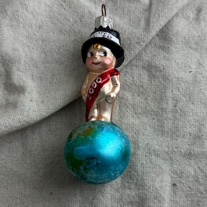 May include: Vintage Christmas ornament featuring a cherubic figure wearing a black hat with "Happy Year" text and a red sash with "2000" text. The figure stands on a blue and green globe. A festive holiday decoration.