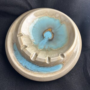 May include: A round ceramic ashtray featuring a beige base glaze with a prominent turquoise blue crystalline glaze effect in the center and along the outer rim. The piece includes four evenly spaced indentations along the inner raised edge.